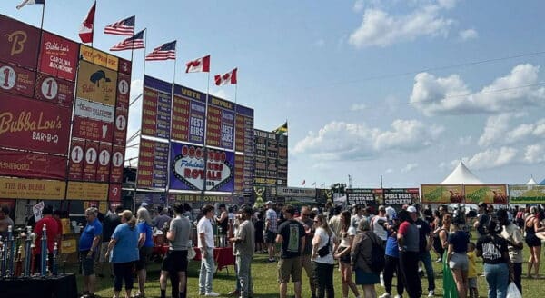A lively outdoor event scene featuring a large crowd of people gathered in front of colorful food vendor signs, with several flags waving in the background under a sunny sky. The atmosphere is bustling and festive, showcasing a community gathering.