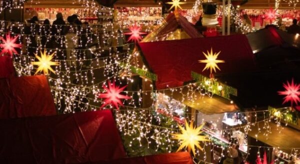 A vibrant market scene illuminated by colorful lights and stars, featuring festive decorations on wooden stalls and rooftops. The atmosphere is lively, suggesting a celebration or holiday event.