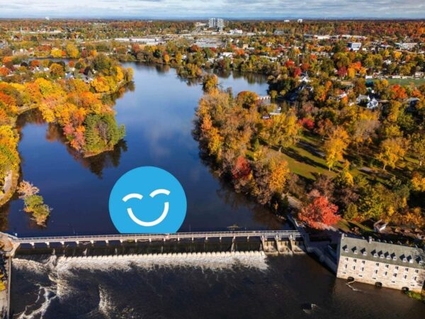 Aerial view of a river with colorful autumn trees lining the shore. A dam is visible on the left, and a blue smiley face graphic is superimposed in the center. The background features a town and a clear sky.