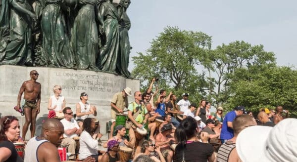 A lively gathering of diverse people enjoying music and dance around a public monument, with musicians playing drums and spectators engaging joyfully. Trees provide shade in the background on a sunny day.