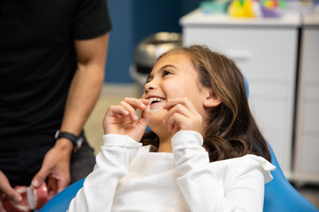 A young girl smiles while sitting in a dental chair, engaged in a playful moment with a dental professional. The scene captures a cheerful atmosphere, promoting a positive dental experience. In the background, bright colors and dental tools are subtly visible, creating a welcoming environment.
