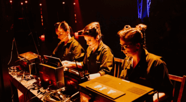 Three individuals work diligently at a table with computers and equipment, illuminated by dramatic lighting. They are focused, likely engaged in a technical or performance-related task within a vibrant event environment.