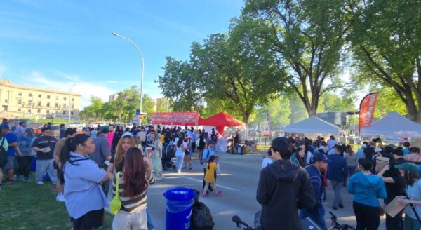 A bustling outdoor event with large crowds gathered around food stalls and tents. Trees line the area under a clear blue sky, creating a lively atmosphere filled with families and individuals enjoying the festivities.