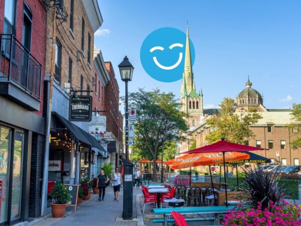 A vibrant street scene features outdoor seating with colorful umbrellas, lined with shops and greenery. In the background, a tall church steeple rises against a clear blue sky, adding to the lively atmosphere.
