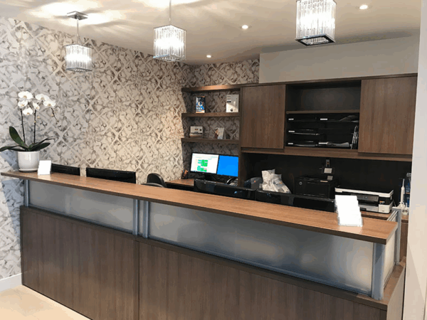 A modern office reception desk with wood finishes, decorative wallpaper, and pendant lights. The desk has computers, printers, and an orchid plant.