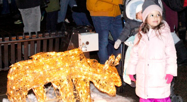 A young girl in a pink coat stands next to a glowing, decorated deer sculpture in a snowy setting, surrounded by people celebrating the festive season.