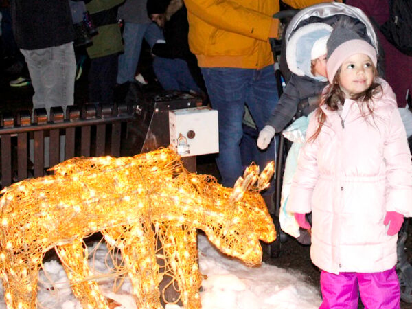 A young girl in a pink coat stands next to a glowing, decorated deer sculpture in a snowy setting, surrounded by people celebrating the festive season.