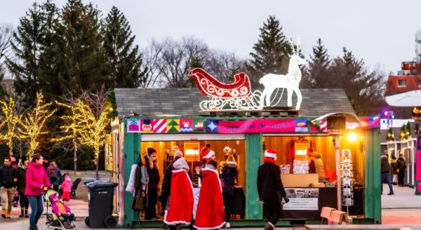 A festive market stall decorated with colorful lights, featuring a sleigh and a horse silhouette. People in Santa costumes gather outside, and families stroll nearby, enjoying the holiday atmosphere among decorated trees.