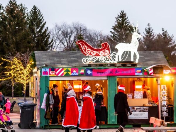 A festive market stall decorated with colorful lights, featuring a sleigh and a horse silhouette. People in Santa costumes gather outside, and families stroll nearby, enjoying the holiday atmosphere among decorated trees.