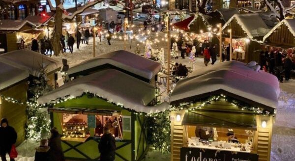 A festive winter market scene with snow-covered stalls adorned with lights. People gather around, exploring various vendors while holiday decorations create a cheerful atmosphere. The setting exudes a warm, communal spirit typical of seasonal celebrations.