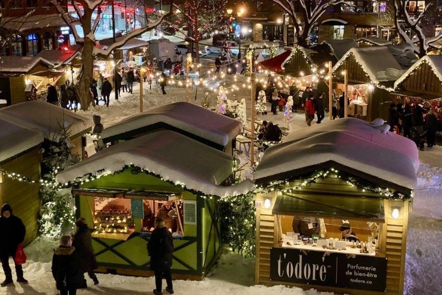 A festive winter market scene with snow-covered stalls adorned with lights. People gather around, exploring various vendors while holiday decorations create a cheerful atmosphere. The setting exudes a warm, communal spirit typical of seasonal celebrations.