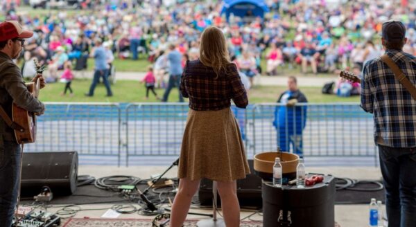 A singer stands on stage with her back to the audience, performing at an outdoor event. Two musicians are visible beside her, and a large crowd gathers in the background, enjoying the performance. A festive atmosphere is evident with people sitting on the grass.