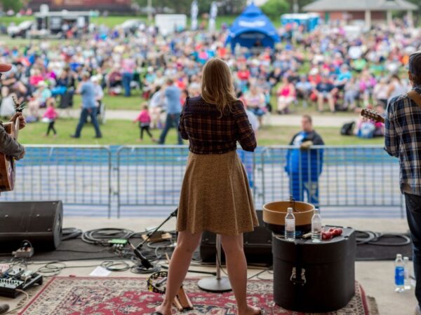 A singer stands on stage with her back to the audience, performing at an outdoor event. Two musicians are visible beside her, and a large crowd gathers in the background, enjoying the performance. A festive atmosphere is evident with people sitting on the grass.