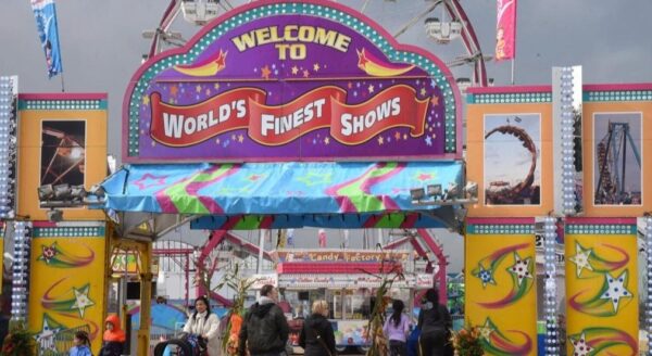 A vibrant entrance archway to a fairground, featuring colorful decorations and the text "Welcome to World's Finest Shows." The scene shows people entering, with amusement rides visible in the background under a cloudy sky.