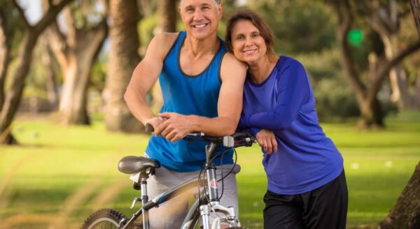 A man and a woman smile together outdoors, leaning against a bicycle. The man wears a blue tank top and shorts, while the woman is dressed in a blue long-sleeve shirt and black pants. The background features green trees and a sunny atmosphere.