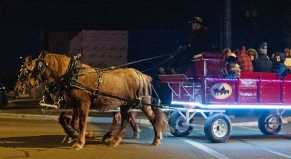 A horse-drawn sleigh with a bright red carriage glides through a nighttime street, carrying several passengers bundled in winter clothing. The sleigh features a bat logo and is illuminated with lights, adding a festive touch to the scene.