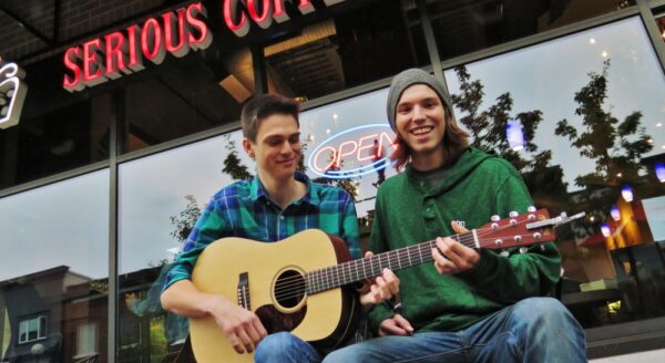 Two young men sit outside a coffee shop, one playing an acoustic guitar while both smile and enjoy the moment. The shop's sign reads "SERIOUS COFFEE," and an "OPEN" sign is visible in the background.