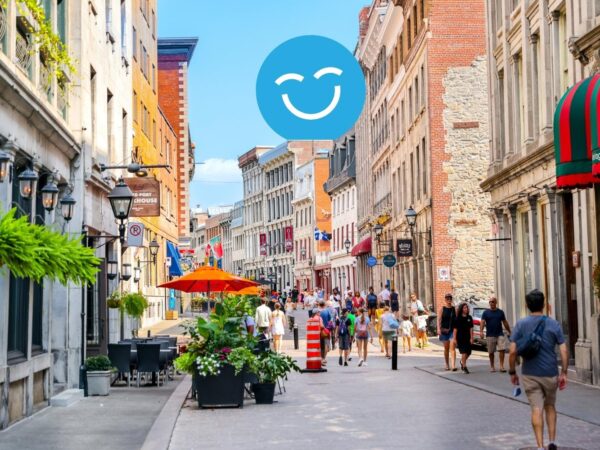 A busy, vibrant street scene with historic buildings, colorful awnings, and pedestrians enjoying a sunny day. A smiling face emoji is superimposed above the street.