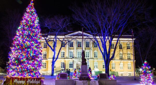 A brightly lit Christmas tree adorned with colorful lights stands in front of a historical building, with snow on the ground. The scene is festive and inviting, showcasing holiday decorations and a warm ambiance at night.