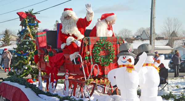 Two Santa Claus figures wave from a festive sleigh adorned with greenery and decorations, accompanied by cheerful snowman figures in a snowy parade setting.