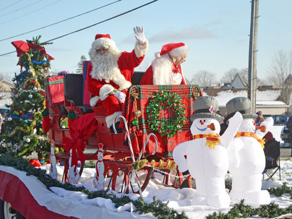 Two Santa Claus figures wave from a festive sleigh adorned with greenery and decorations, accompanied by cheerful snowman figures in a snowy parade setting.