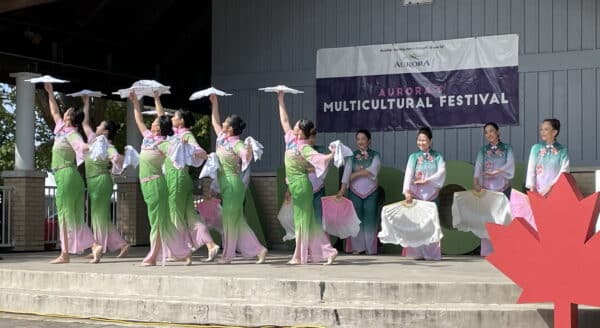 A group of performers in colorful traditional costumes dances on stage at a multicultural festival. They hold umbrellas and exhibit coordinated movements, celebrating cultural diversity. A large Canadian maple leaf decoration is visible in the foreground, enhancing the festive atmosphere.