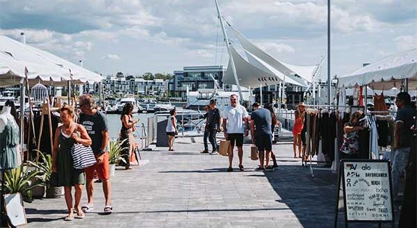 A bustling waterfront market with people browsing stalls under white tents, surrounded by boats and a scenic marina. Bright skies create a lively atmosphere as visitors enjoy shopping and exploring the area.