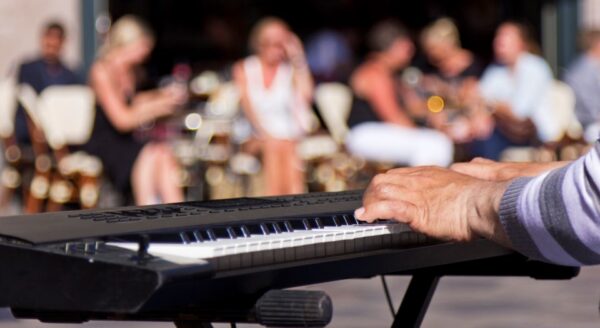 A close-up view of a person's hands playing a keyboard, with blurred figures of people enjoying a casual outdoor gathering in the background.
