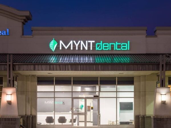 The image shows the exterior of a modern dental clinic named MYNT dental, illuminated at dusk. The building features large windows and a prominent sign with the clinic's name, indicating its location within a shopping plaza.