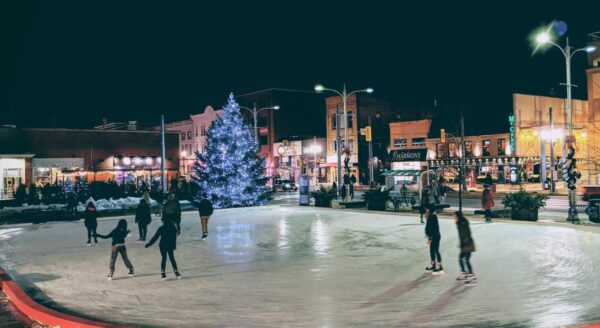 A festive outdoor ice skating rink at night, surrounded by illuminated buildings and a large, decorated Christmas tree. Skaters enjoy the icy surface, creating a joyful winter atmosphere.