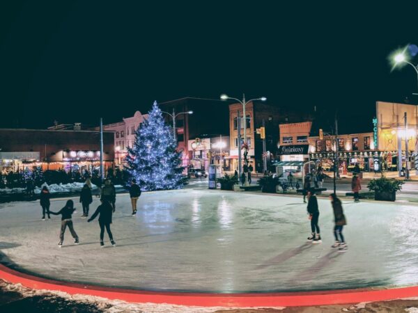 A festive outdoor ice skating rink at night, surrounded by illuminated buildings and a large, decorated Christmas tree. Skaters enjoy the icy surface, creating a joyful winter atmosphere.