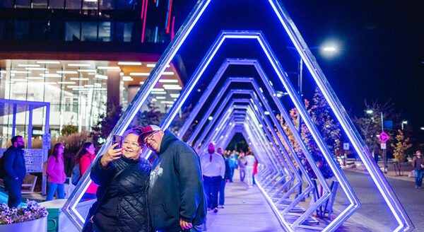 A couple poses for a selfie in a brightly lit pathway featuring neon blue triangular arches. People stroll in the background at night, showcasing a lively urban atmosphere.