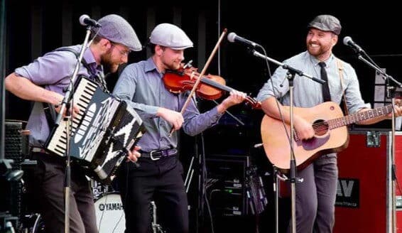 Three musicians perform on stage, playing an accordion, violin, and guitar. They are dressed in grey outfits and flat caps, creating a lively and engaging atmosphere.