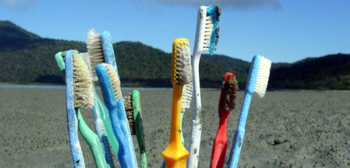 A collection of colorful toothbrushes is positioned upright on a sandy surface, surrounded by a natural landscape of hills and a blue sky in the background, highlighting environmental concerns related to plastic waste.