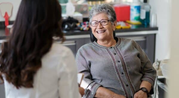 A smiling older woman sits in a dental office, engaging with a dental professional. The setting is bright and welcoming, reflecting a friendly atmosphere. The 123Dentist Smiley adds a cheerful touch to the scene, promoting a positive dental experience.