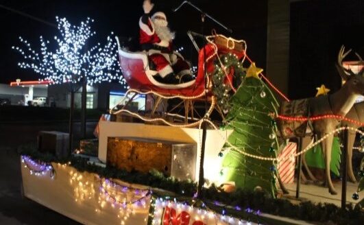 A festive holiday parade float featuring Santa Claus in a red sleigh, surrounded by a decorated Christmas tree and illuminated by colorful lights. The scene is set against a night backdrop, adding to the cheerful, holiday atmosphere.