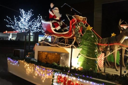 A festive holiday parade float featuring Santa Claus in a red sleigh, surrounded by a decorated Christmas tree and illuminated by colorful lights. The scene is set against a night backdrop, adding to the cheerful, holiday atmosphere.