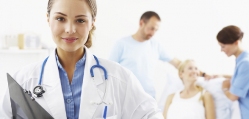 A female doctor in a white coat holds a clipboard, smiling at the camera. In the background, a patient is being attended to by a male nurse and another healthcare professional, depicting a caring medical environment.
