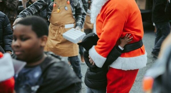A festive scene with Santa Claus in a red suit warmly embracing a child, surrounded by festive celebrations and people. Others nearby are engaged in holiday activities, creating a joyful atmosphere.