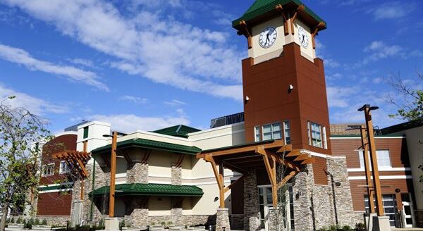 A modern building featuring a clock tower and a green roof, complemented by stone and wooden accents. The structure is set against a bright blue sky with scattered clouds, showcasing contemporary architectural design.