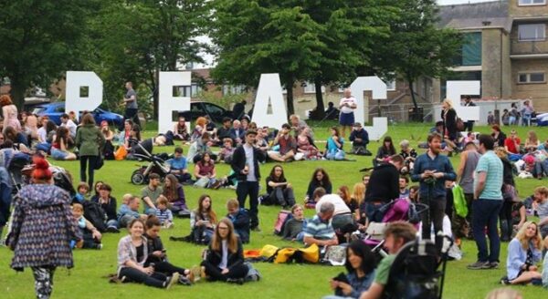 A crowd of people sitting on a grassy field, some enjoying a picnic, with large white letters spelling "PEACE" in the background. Trees and buildings are visible, creating a lively, community atmosphere.
