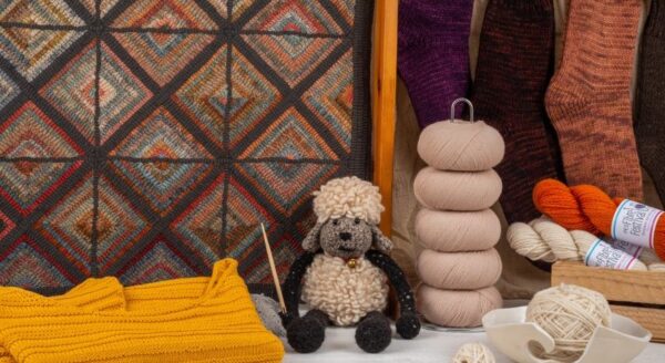 A cozy display featuring a handmade stuffed sheepdog surrounded by colorful yarn spools, a knitted sweater, and various knitted socks against a patterned backdrop.