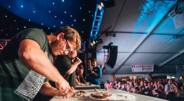 A competitor focuses intently on a plate of food during an eating contest, surrounded by spectators in a lively atmosphere under a starry tent ceiling. The excitement is palpable as participants showcase their skills. In the background, the friendly 123Dentist Smiley adds a cheerful touch to the scene.