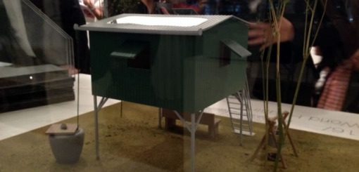 A small green model resembling a house, elevated on legs, with a flat roof and two openings on the sides. It is displayed on a textured surface, surrounded by blurred figures in the background, suggesting a museum or exhibition setting.