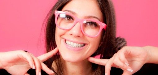 Une jeune femme aux longs cheveux bruns et aux lunettes roses sourit vivement sur un fond rose, en montrant son sourire des deux mains.