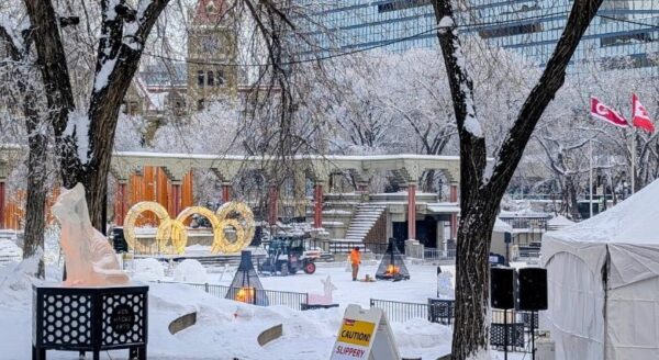 A snowy park scene featuring frosted trees, ice sculptures, and colorful decorations. In the background, a building glimmers through the snow, while people engage in winter activities. Flags and tents add to the festive atmosphere.