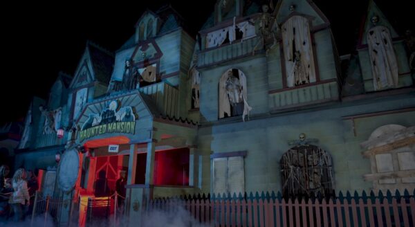 A spooky haunted house attraction features eerie lighting, creepy decorations, and a foggy atmosphere, inviting visitors to experience frights and thrills. The entrance is marked with a sign, and ghostly figures can be seen in the shadows.