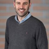 A smiling man with a beard stands in front of a brick wall. He is wearing a dark sweater over a light blue collared shirt.