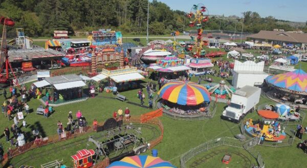 A vibrant carnival scene featuring colorful rides, game booths, and tents. Families and children enjoy the attractions on a sunny day, surrounded by green grass and trees in the background.