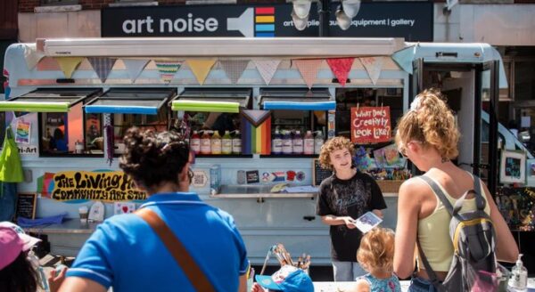A vibrant outdoor scene at a market featuring a colorful food truck. People are gathered around, with one vendor engaging with customers. Banners and artwork enhance the lively atmosphere, showcasing a community event.